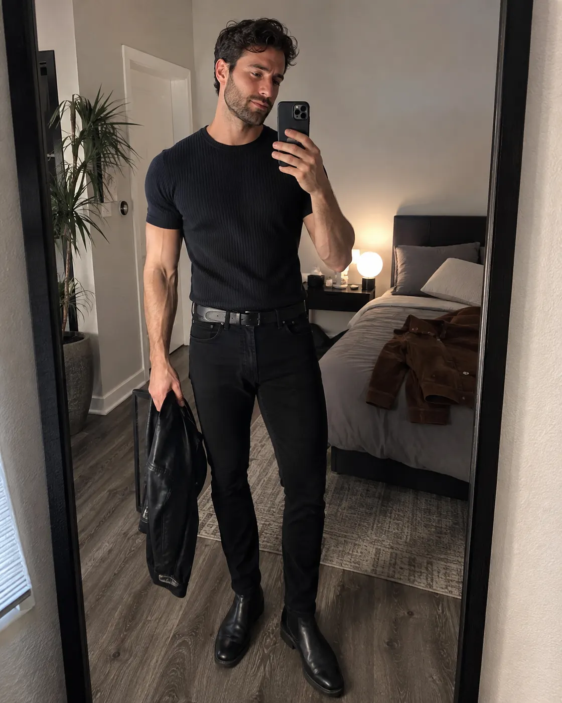 A man in a fitted black t-shirt, black jeans and black boots holding a leather jacket.