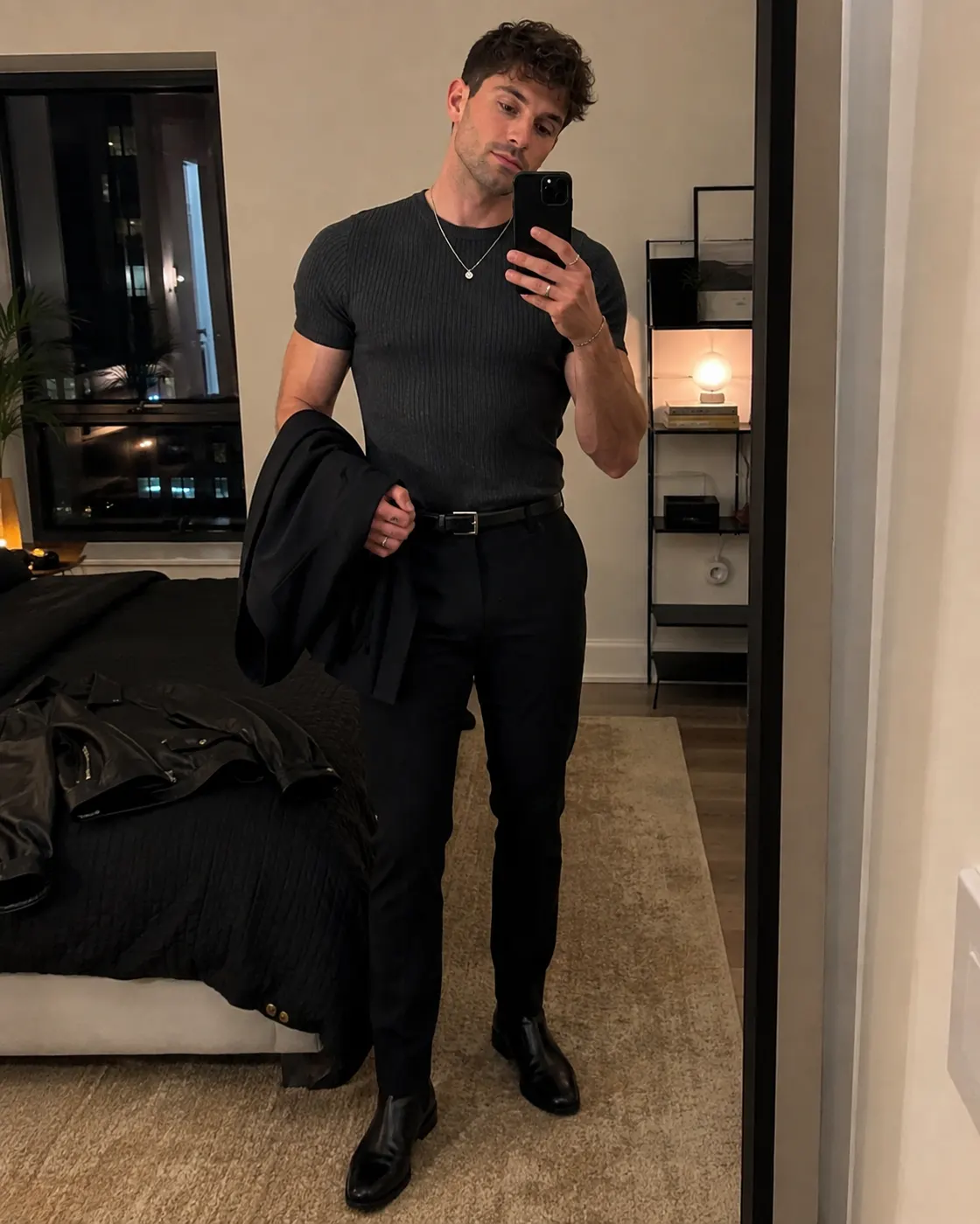 A man in a fitted black t-shirt and black trousers holding a jacket, city lights behind him.