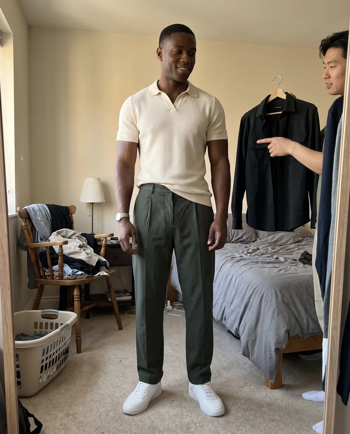 A man in a cream knitted polo and olive trousers while a friend holds up a black shirt on a hanger.