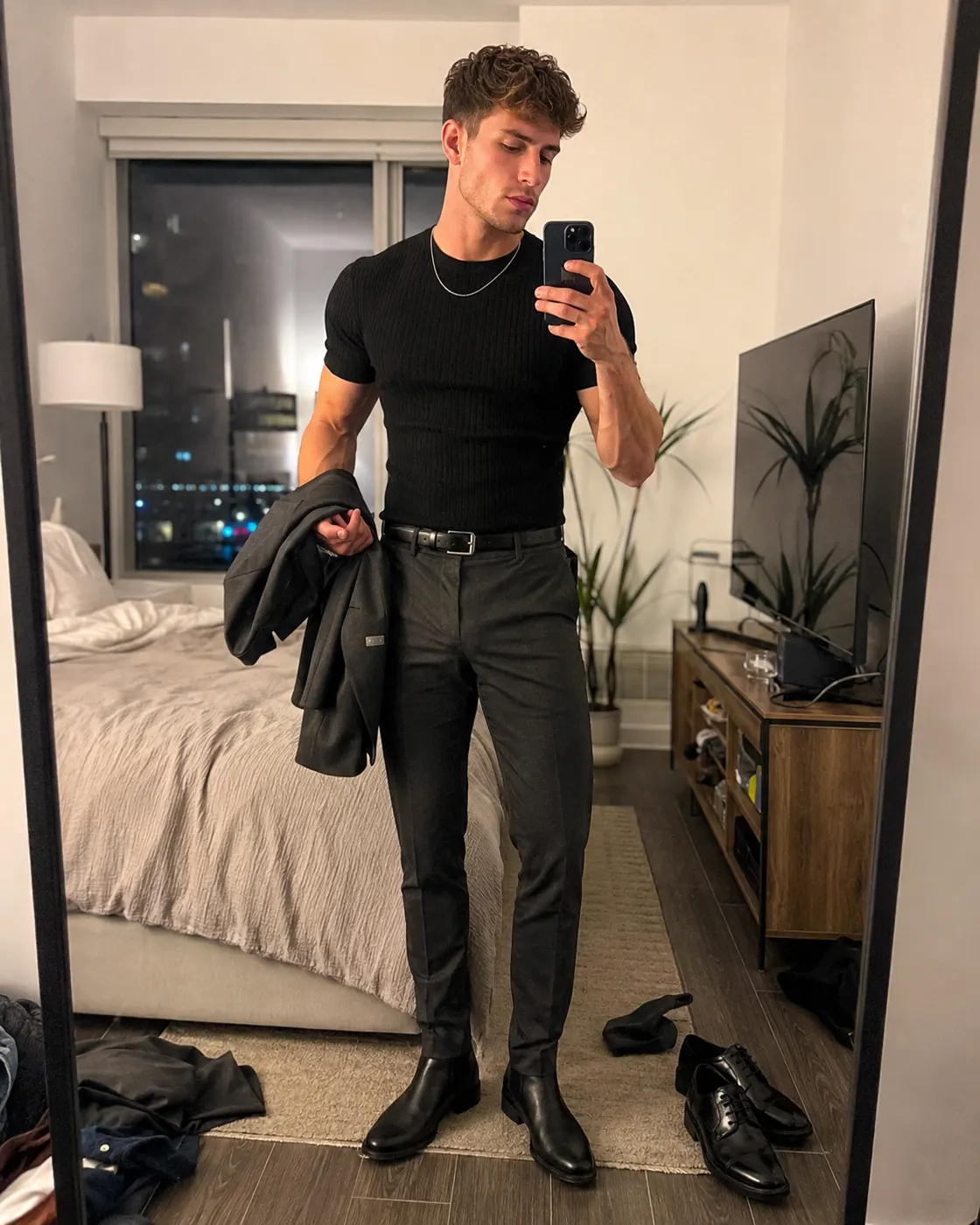 A man in a fitted black t-shirt and olive trousers with black boots and brown loafers side by side on the floor.