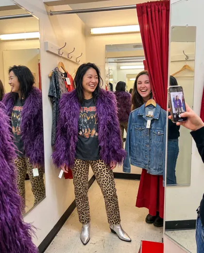 A woman in a purple faux-fur coat, band t-shirt, leopard-print trousers and silver boots in a fitting room while two friends watch.