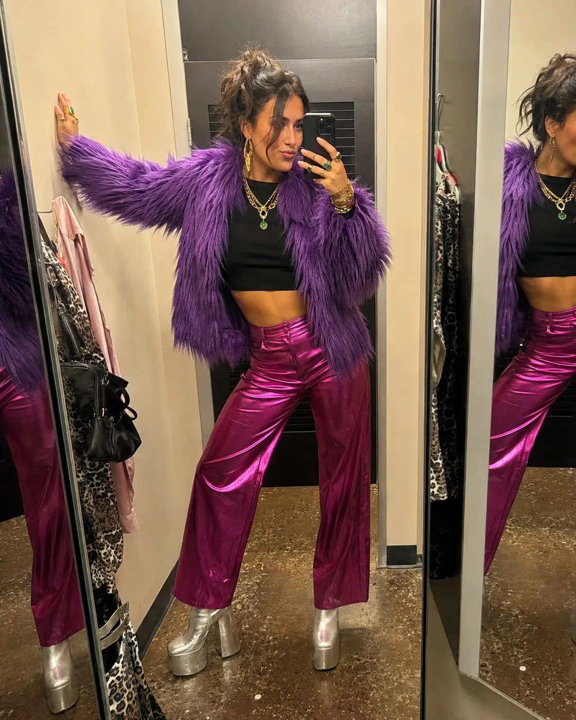 A woman in a purple faux-fur jacket, magenta metallic trousers and silver platform boots in a fitting room.