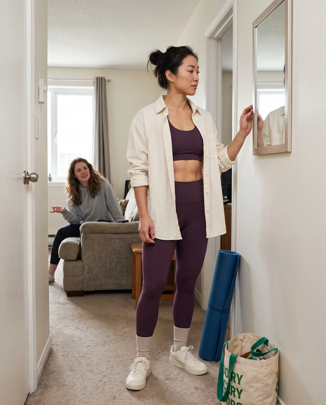 A woman in leggings and a cropped top with an open cream overshirt, checking a mirror in an apartment hallway.