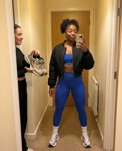 A woman in a cobalt gym set and black bomber taking a hallway mirror photo while a friend holds up two pairs of trainers.