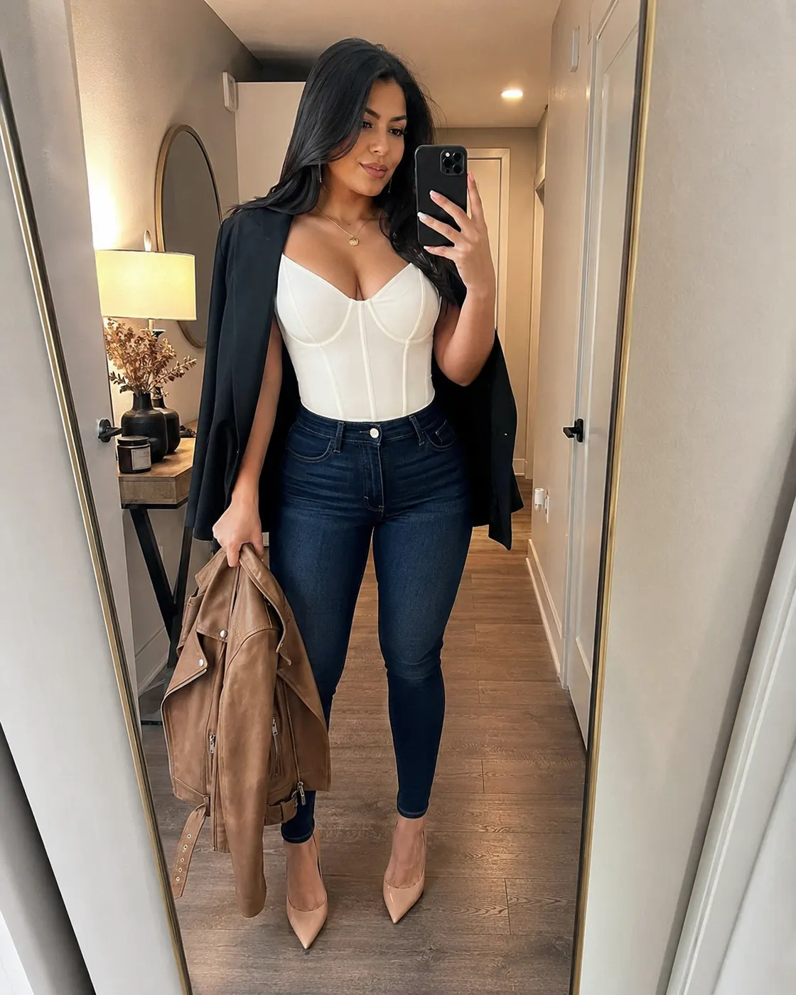 A woman in a white corset top and dark jeans holding a tan suede jacket, nude heels on.