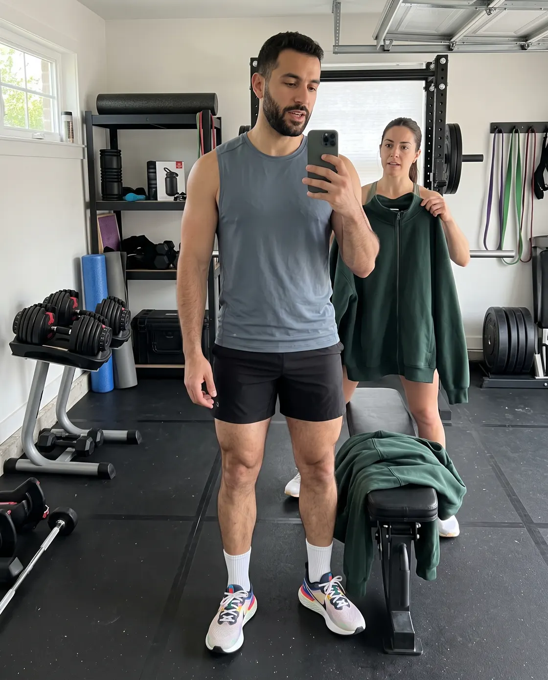 A man in a grey training top and shorts photographing his reflection in a home gym while a friend holds up a green sweatshirt.