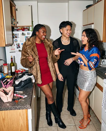 Three friends in a small kitchen before going out: a red dress under a fur coat, an all-black suit, and a sequin skirt with a sports jersey.
