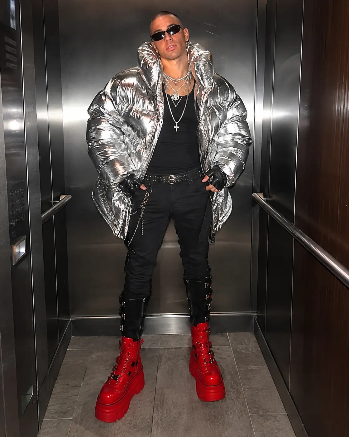 A man in a metallic silver puffer jacket, black cargo trousers and red platform boots in a lift.