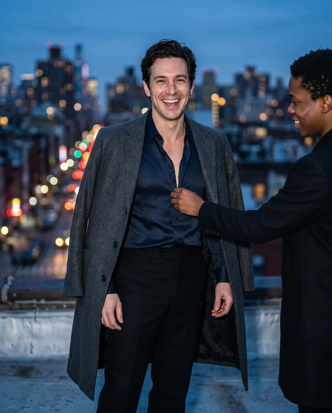 A man in a long grey coat over a dark shirt on a balcony at night, a friend adjusting his lapel.