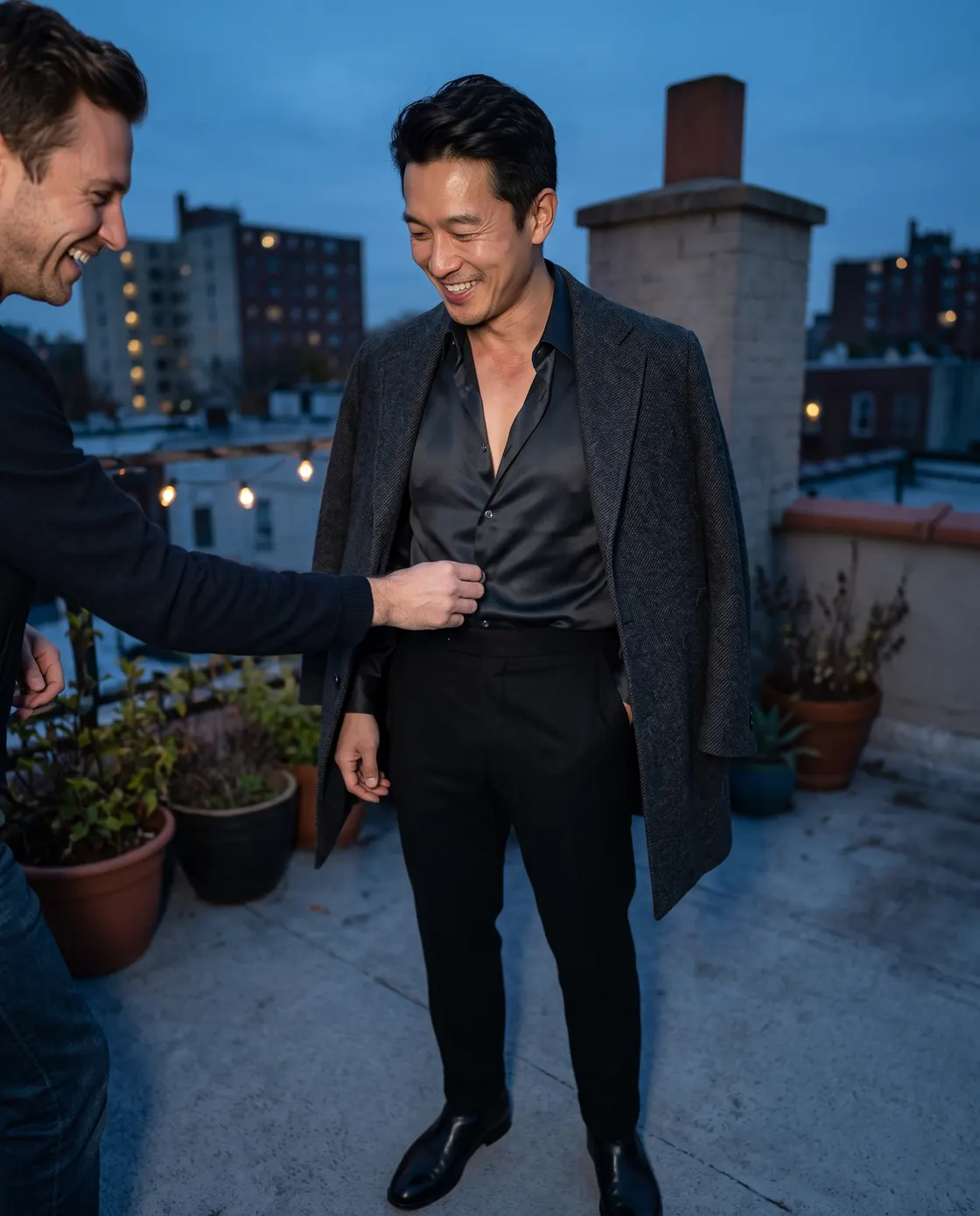 A man in a black shirt and dark trousers on a rooftop at dusk while a friend adjusts his collar.