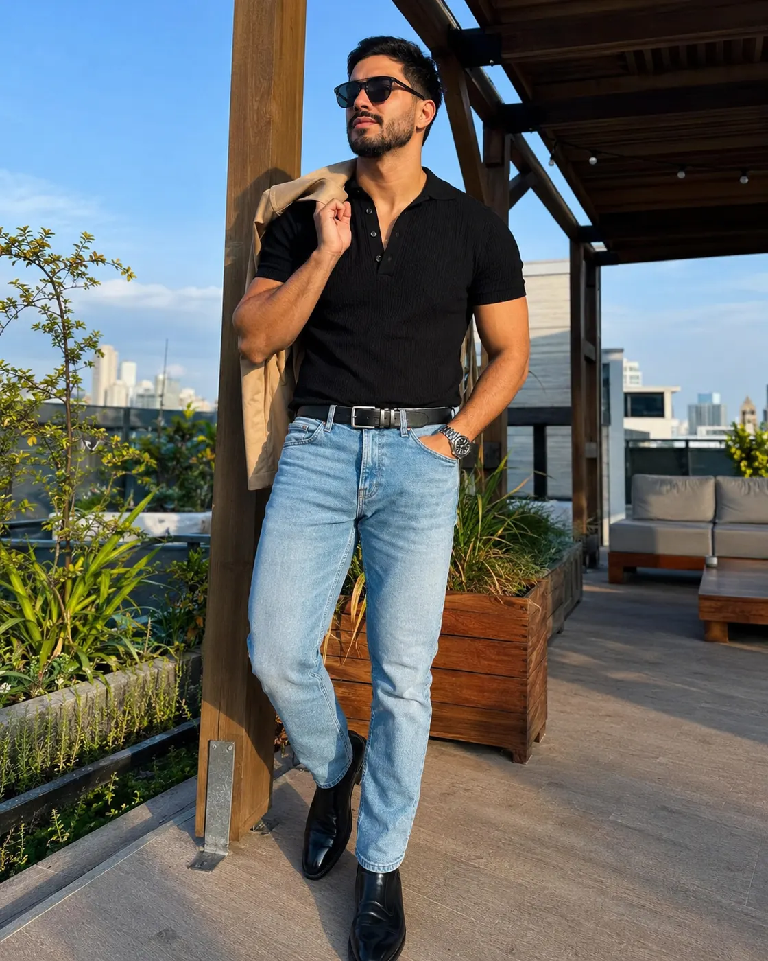 A man in a black polo, light jeans and black boots on a rooftop terrace, jacket over one shoulder.