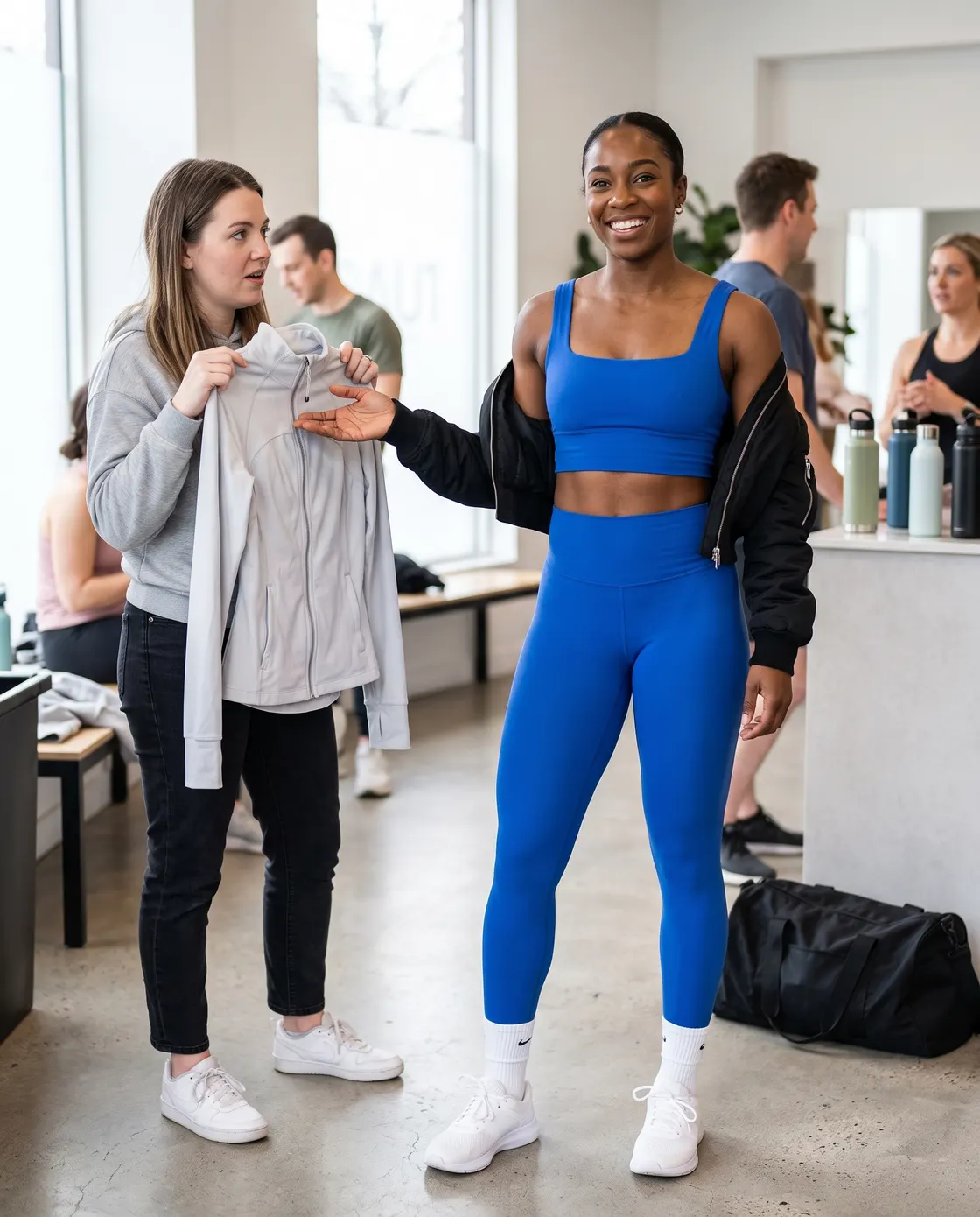 A woman in a bright blue gym set and bomber jacket in a studio while a friend holds up a grey hoodie.